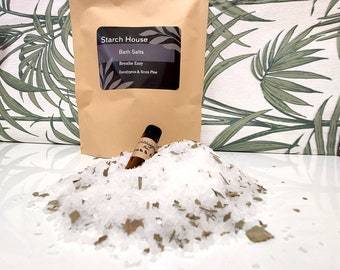May include: A brown paper bag with a label that reads "Starch House Bath Salts Breathe Easy Eucalyptus & Scots Pine". A small brown glass bottle with a white label is sitting on a pile of white bath salts with green leaves.