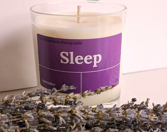 May include: A purple and white soy candle with the word "Sleep" and the scent "Lavender" printed on the label. The candle is in a clear glass jar and is surrounded by dried lavender flowers.