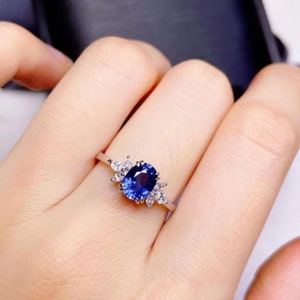 Può includere: Un anello d'argento con una grande pietra preziosa blu scuro di taglio ovale come elemento centrale. L'anello è impreziosito da pietre preziose chiare più piccole a forma di pera su entrambi i lati della pietra principale, creando un design a grappolo. L'anello è mostrato su un dito.