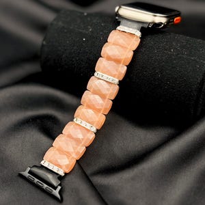 May include: A peach-colored watch band with a faceted design and silver accents. The band is made of rectangular beads and is attached to a silver watch face with a black screen. The band is displayed on a black surface.