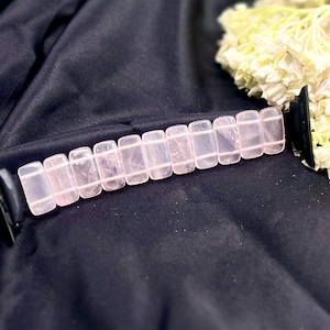 May include: A rose quartz bracelet with rectangular beads. The light pink stones are strung together, resting on a dark, textured surface. A cluster of white flowers is in the background.
