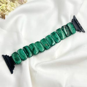 Op de afbeelding: Een horlogeband met rechthoekige groene malachietstenen. De stenen zijn verbonden door een zwarte band. De malachiet heeft een kenmerkend patroon van wervelende groentinten. De horlogeband wordt gepresenteerd op een witte, getextureerde ondergrond.