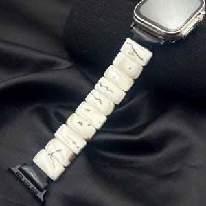 May include: A smartwatch featuring a rectangular metallic case and a watch band composed of rectangular white beads with brown veining. The band connects to the watch via black adapters, and the watch face is dark and reflective.