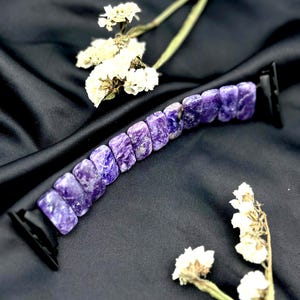 May include: A watch strap made of purple stone with a black clasp. The strap features rectangular, polished stones in shades of purple and white. It is displayed on a black satin surface, with white flowers in the background.
