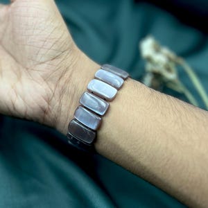 Op de afbeelding: Een armband met rechthoekige, iriserende, grijstinten kralen. De armband wordt om een pols gedragen, waarbij de kralen licht reflecteren en een reeks grijstinten weergeven. Het ontwerp van de armband is eenvoudig en elegant, geschikt voor dagelijks gebruik.