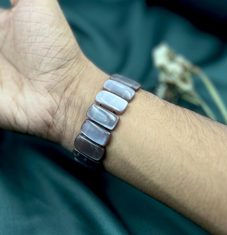 Op de afbeelding: Een armband met rechthoekige, iriserende, grijstinten kralen. De armband wordt om een pols gedragen, waarbij de kralen licht reflecteren en een reeks grijstinten weergeven. Het ontwerp van de armband is eenvoudig en elegant, geschikt voor dagelijks gebruik.