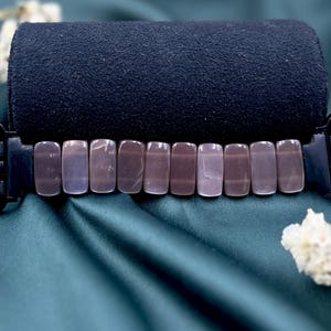 Op de afbeelding: Een zwarte Apple Watch-band met rechthoekige, gepolijste, mauvekleurige stenen. De band wordt getoond op een donkerblauwe, opgerolde stof tegen een teal-satijnen achtergrond, met witte bloemaccenten.