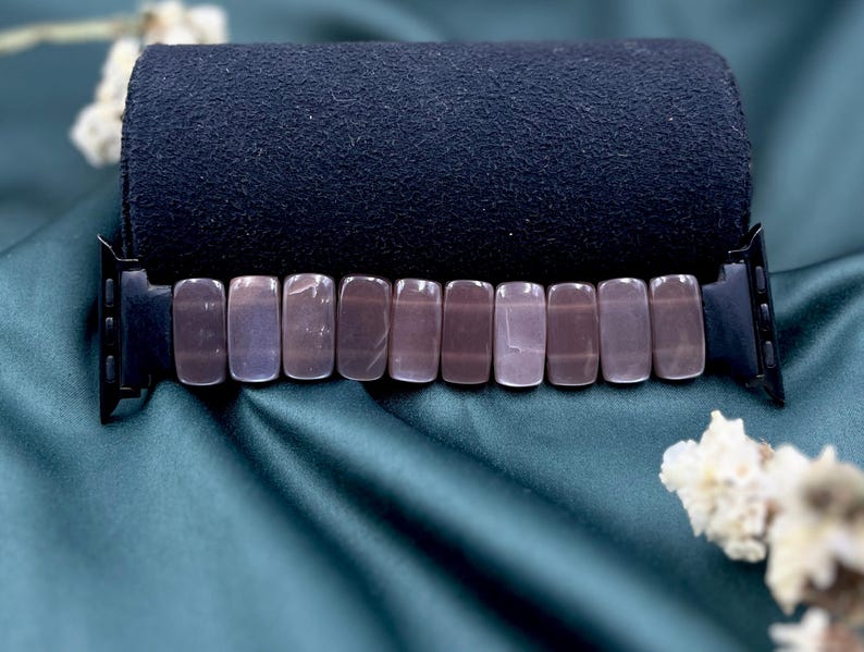 Op de afbeelding: Een zwarte Apple Watch-band met rechthoekige, gepolijste, mauvekleurige stenen. De band wordt getoond op een donkerblauwe, opgerolde stof tegen een teal-satijnen achtergrond, met witte bloemaccenten.
