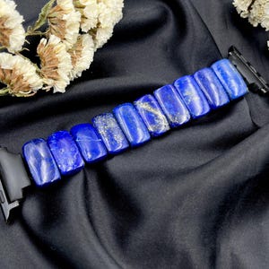 Pode incluir: Uma pulseira de relógio azul escuro feita de pedras de lápis-lazúli polidas. As pedras retangulares têm manchas douradas. A pulseira é presa a conectores pretos. O fundo é um tecido preto com flores brancas secas.