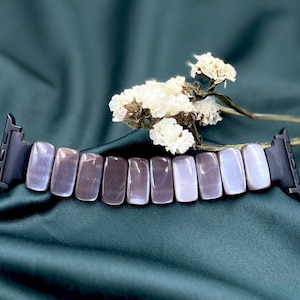 May include: A watch band with rectangular, iridescent, gray-purple stones. The band has black connectors on each end. The background is a dark teal fabric, and a sprig of white flowers is in the upper right corner.