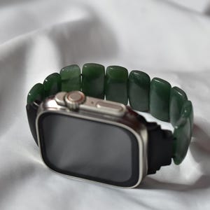 Może przedstawiać: Srebrny smartwatch z czarnym paskiem i bransoletką z zielonych kamieni jadeitu. Tarcza zegarka jest ciemna, a kamienie jadeitu są prostokątne i wypolerowane. Zegarek jest umieszczony na białym, teksturowanym tle.