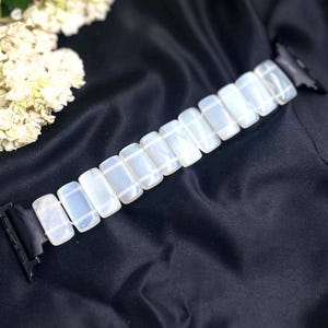May include: A white rectangular link watch band with black connectors. The band is made of a series of white, translucent, rectangular links. The band is displayed on a black, silky fabric, with white flowers in the background.