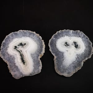 May include: Two white agate slices with a dark gray, almost black, center. The agate slices are translucent and have a layered, banded appearance.
