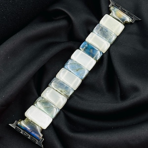 May include: An Apple Watch band featuring a rectangular design with alternating white and blue-grey stone segments. The band has black connectors and is displayed on a black satin background. The stones have a marbled appearance.