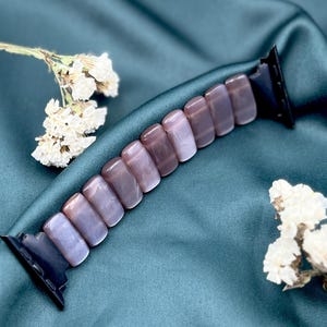 Op de afbeelding: Een horlogebandje gemaakt van rechthoekige, gepolijste, mauvekleurige stenen. De band is bevestigd aan zwarte connectoren. De achtergrond is een teal-kleurige satijnen stof, met witte gedroogde bloemen op de voorgrond.