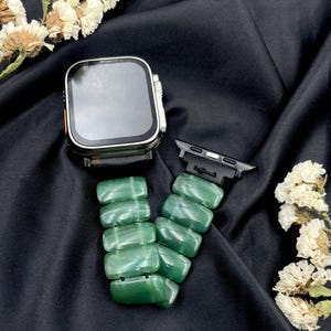 May include: A silver smartwatch with a black strap and a green stone watch strap. The green strap is made of rectangular, polished stones. The watch and strap are displayed on black fabric with white flowers.