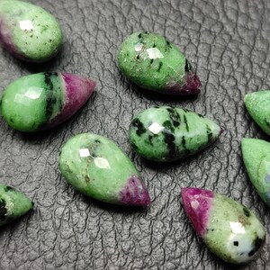 May include: A collection of ten teardrop-shaped green and pink gemstones. The stones have a faceted cut and are arranged on a textured black surface.