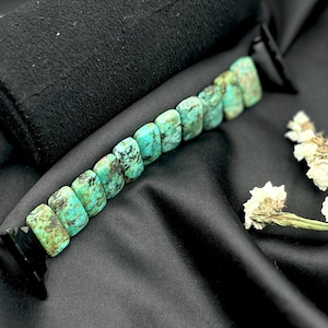 May include: A turquoise bracelet with rectangular beads. The bracelet is displayed on a black surface with a black object and white flowers in the background. The turquoise beads have a mix of green and brown colors.