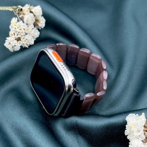 Op de afbeelding: Een zilveren smartwatch met een zwart scherm en een oranje knop. Het is gekoppeld aan een bruine, kralen armband. De achtergrond is een donkergroene stof met witte bloemen.
