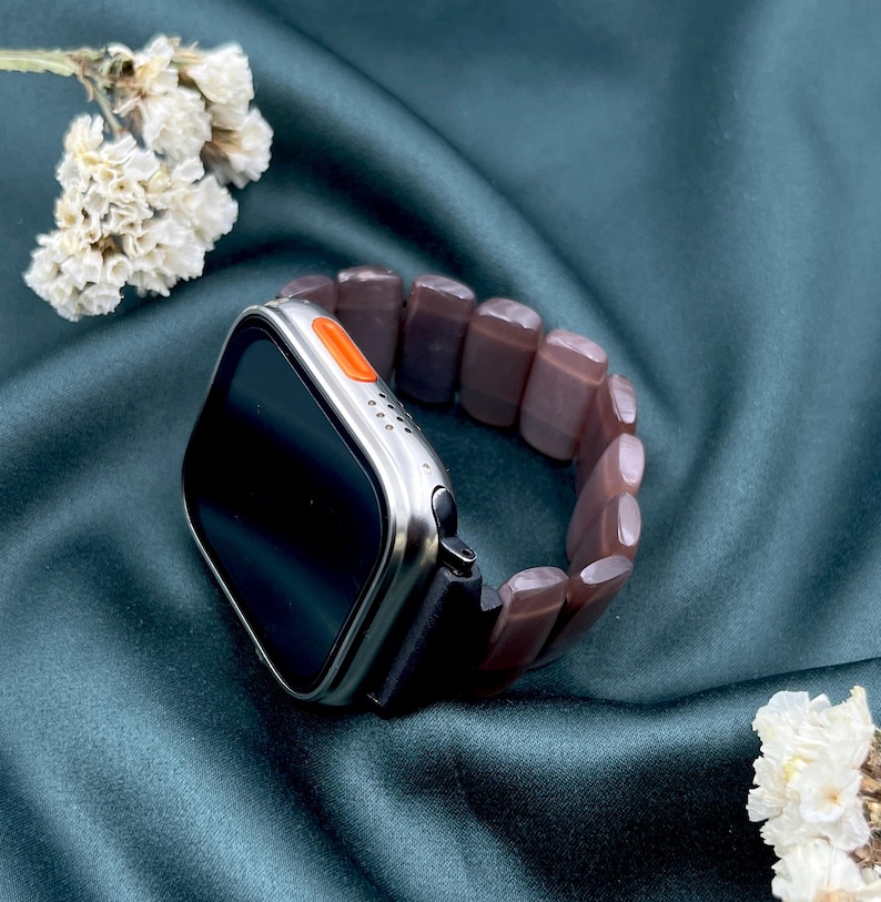Op de afbeelding: Een zilveren smartwatch met een zwart scherm en een oranje knop. Het is gekoppeld aan een bruine, kralen armband. De achtergrond is een donkergroene stof met witte bloemen.