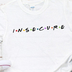 May include: White t-shirt with the word "INSECURE" in colorful letters, styled like the TV show "Friends." The shirt is paired with white sneakers and blue jeans, creating a casual, everyday look.