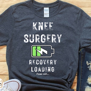 May include: A dark gray t-shirt with the text "Knee Surgery Recovery Loading Please wait..." and a green battery icon with a lightning bolt.