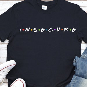 May include: A black t-shirt with the word "INSECURE" written in white letters with colorful dots between each letter.