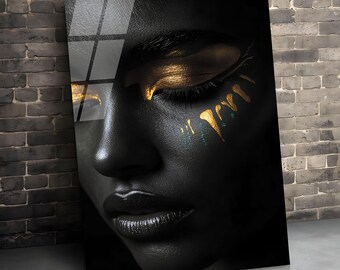 May include: A close-up of a black portrait with gold accents. The subject's face is painted black, with gold eyeshadow and gold paint strokes on the cheek. The lips are dark, and the image is displayed on a glass panel.