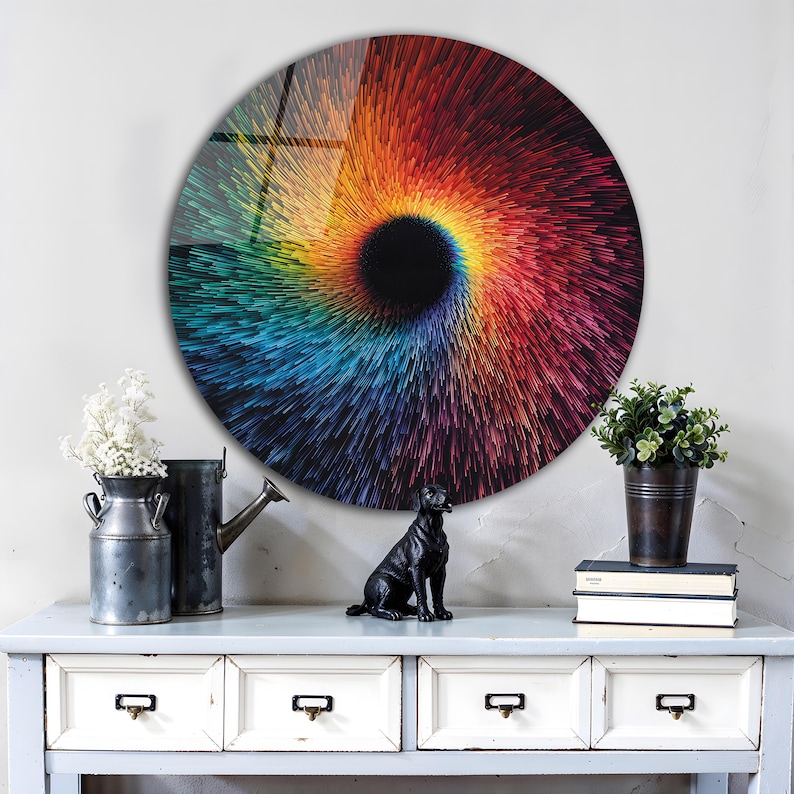 May include: A round wall art piece with a vibrant, colorful spiral design. The artwork features a black center that radiates outward with streaks of red, orange, yellow, green, and blue. The art is displayed above a white dresser with three drawers.