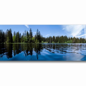 May include: A panoramic view of a tranquil lake surrounded by a dense forest of evergreen trees. The water is a deep blue and reflects the sky and trees above. The sky is a bright blue with wispy white clouds.