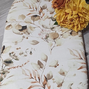 May include: An ironing board cover featuring a botanical leaf design in brown, beige, and green hues. The cover is decorated with artificial orange and yellow flowers, enhancing the ironing board's aesthetic appeal.