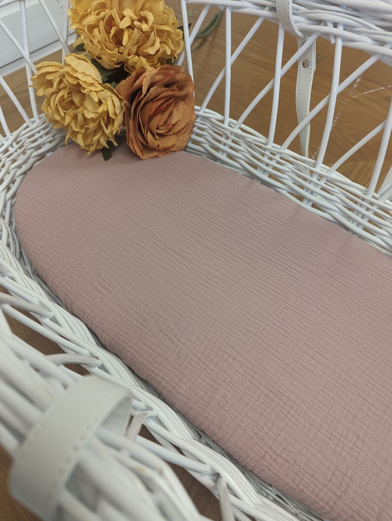 May include: A white wicker basket with a pink fitted sheet inside. The basket is empty and has a handle on the top. There are three yellow and orange silk flowers on top of the basket.