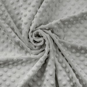 Op de afbeelding: Close-up van een zachte, grijze, textuurrijke stof met een reliëfpatroon van stippen. De stof is gevouwen en gedraaid, waardoor de textuur en zachtheid zichtbaar worden.