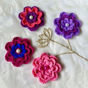 May include: Four crocheted flowers with pearl centres. The flowers are pink, purple and magenta. The flowers are arranged on a white fabric background.
