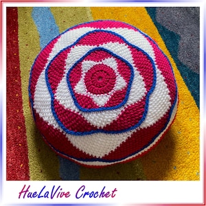 May include: A crocheted round cushion with a pink, white, and blue geometric pattern. The cushion is on a colorful rug.