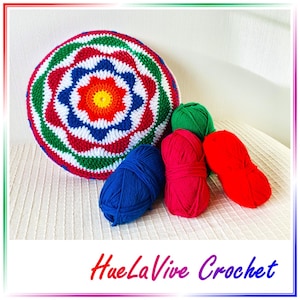 May include: A colorful crocheted round pillow with a flower design in the center. The pillow is surrounded by balls of yarn in blue, red, and green.