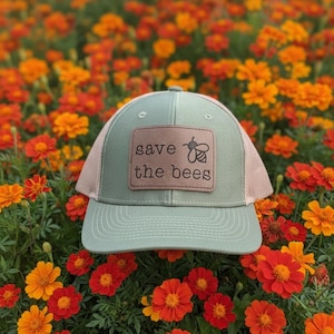 May include: A sage green trucker hat with a tan mesh back and a curved brim. The hat features a brown leather patch with the words "save the bees" and a bee graphic. The hat is set against a backdrop of orange and red flowers.