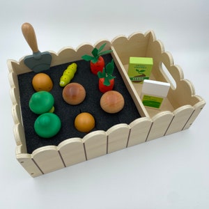 Könnte beinhalten: Ein hölzernes Spielzeug-Gartenset mit einem Zaun, einer grünen Schaufel, einem grünen Wurm, braunen und orangefarbenen Gemüsesorten und zwei Schachteln mit der Aufschrift "Bio-Gemüsesamen" und "NPK-Dünger".