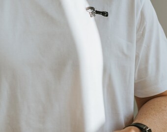 以下が含まれることがあります： 白いTシャツを着て、前面に小さく黒と白のエスプレスポータフィルターのデザインが施されています。その人は、銀色のポータフィルターを手に持っています。