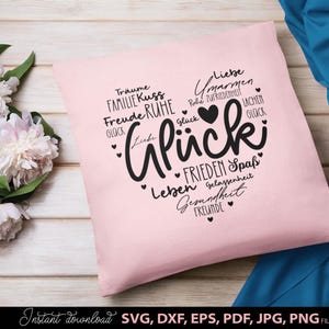 Puede incluir: Un cojín cuadrado rosa claro con un diseño en forma de corazón negro lleno de palabras alemanas como "Glück" y "Liebe". El cojín está sobre una superficie de madera con flores blancas y tela azul en el fondo. Las palabras "Instant download SVG, DXF, EPS, PDF, JPG, PNG FILES" están en la parte inferior.