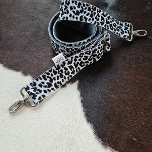 May include: A white and black leopard print adjustable strap with silver hardware. The strap is coiled and ready to be attached to a bag.