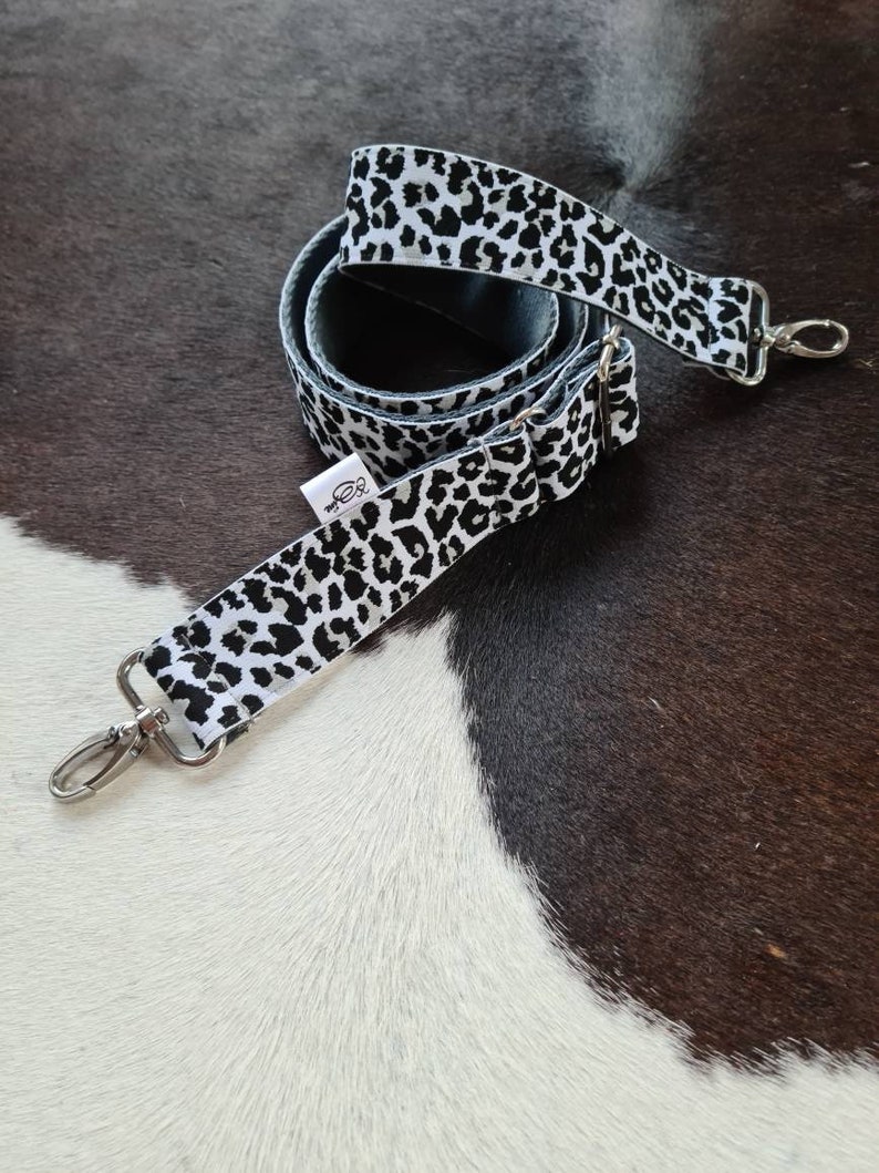 May include: A white and black leopard print adjustable strap with silver hardware. The strap is coiled and ready to be attached to a bag.