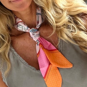Könnte beinhalten: Ein Seidenschal in Rosa, Orange und Weiß. Der Schal hat ein Muster aus Reitausrüstung und Tupfen. Er ist um den Hals gebunden, wobei die Enden herunterhängen.