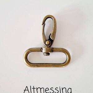 May include: A gold-toned metal clasp with a spring-loaded opening. The clasp is designed for attaching to a strap or chain.