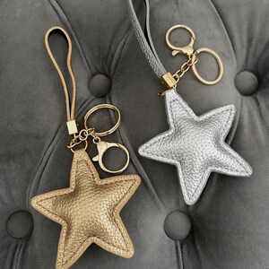 Peut inclure: Deux porte-clés en forme d'étoile. L'un est doré, l'autre argenté, tous deux avec un aspect texturé. Chaque porte-clés a une dragonne assortie et une quincaillerie dorée, comprenant un porte-clés et un fermoir. Les porte-clés sont sur une surface grise capitonnée.