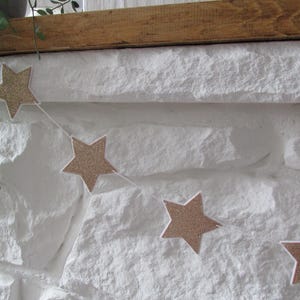May include: A gold glitter star garland with white string. The garland is hanging on a white brick wall.