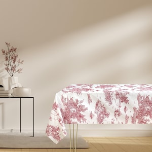 Puede incluir: Un mantel blanco con un patrón floral rojo. El mantel está puesto sobre una mesa con un marco de metal negro. La mesa está en una habitación con una pared blanca y un jarrón con una pequeña rama de árbol.