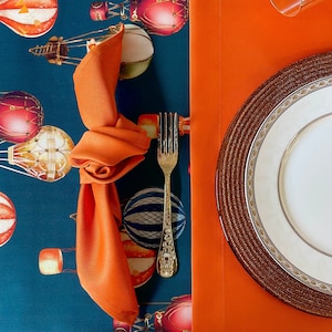May include: A teal tablecloth with a hot air balloon pattern and a brown wicker charger plate with a white dinner plate on top. A silver fork and an orange napkin are on the table.