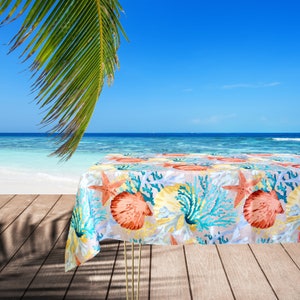 Puede incluir: Una escena de playa tropical con una mesa cubierta con un mantel colorido con un diseño de conchas y corales. El mantel es azul claro con acentos naranja, amarillo y turquesa.