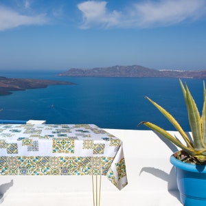 May include: A white tablecloth with a colorful geometric pattern is laid out on a white table with a view of a blue ocean and a distant island. A potted plant with green leaves is in the foreground.